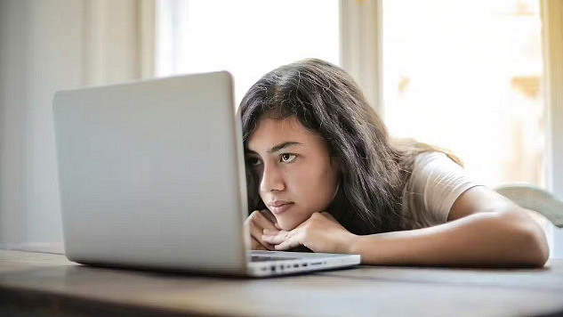 A teenage girl leans her head on a table and looks at a laptop screen.