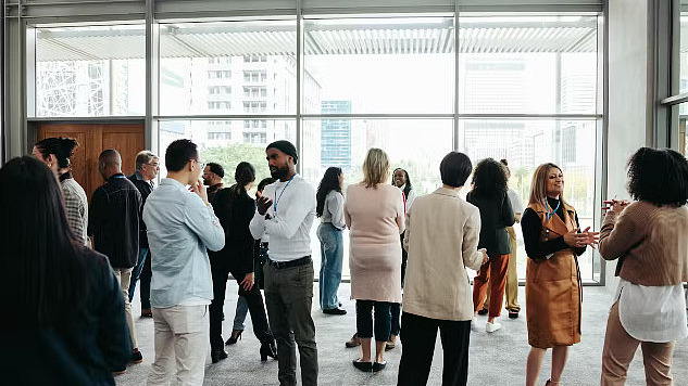 A diverse crowd of people in an office building, standing in small groups having conversations, representing networking in a professional environment.