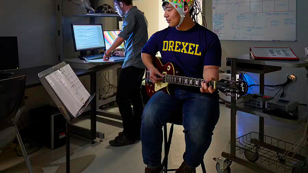 मस्तिष्क की गतिविधि को मापने वाले इलेक्ट्रोड से ढका हेलमेट पहनकर युवक गिटार बजाता है