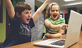 duas crianças na frente de um computador comemorando um sucesso com as mãos para o ar e com grandes sorrisos