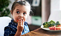 एक छोटी बच्ची टेबल पर बैठकर थाली से सब्जियां खाती है