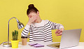 Una joven sentada en un escritorio frente a una pared amarilla, se tapa los ojos con una mano y usa la otra para proteger la pantalla de su computadora, sugiriendo "No quiero mirar esto".
