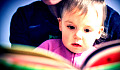 年幼的孩子坐在成人的腿上，看着一本彩色书的书页