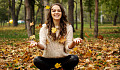 a smiling young woman sitting in the forest amidst falling autumn leaves