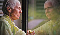 oudere persoon die een appel eet en naar haar spiegelbeeld in een raam kijkt