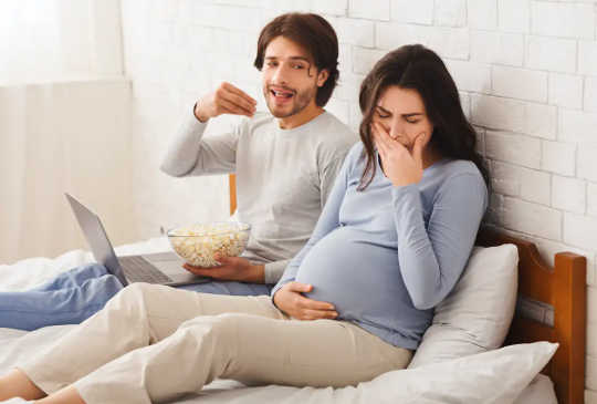 ผู้ชายกำลังกินอาหารบนเตียงในขณะที่สตรีมีครรภ์ปิดจมูก