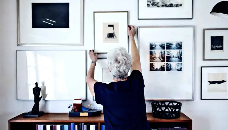 אישה מבוגרת תולה תצלום ממוסגר על קיר מכוסה באמנות ממוסגרת אחרת
