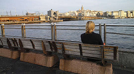 一个女人独自坐在面对水和城市天际线的长凳上