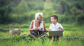 een jong kind met een laptop in gesprek met zijn grootmoeder die buiten zit met een picknickmand
