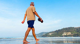 homem andando na praia segurando suas sandálias na mão