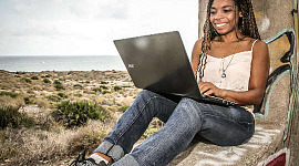 Sırtı ağaca yaslanmış oturan genç bir kadın dizüstü bilgisayarında çalışıyor.