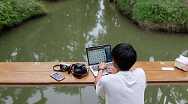 en ung dreng på et skib med sin bærbare computer åben og et kamera og en mobiltelefon ved siden af ​​sig.