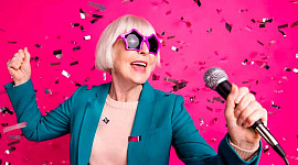 femeie cu părul cărunt, purtând ochelari de soare de un roz funky, cântând ținând un microfon