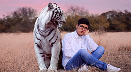 νεαρός άνδρας κάθεται σε ένα χωράφι με μια μεγάλη τίγρη να κάθεται δίπλα του