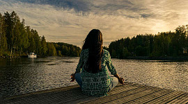kvinne, sett fra baksiden, sittende i lotusstilling på en kai ved en innsjø