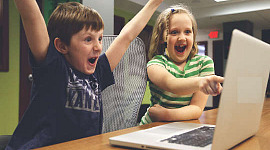doi copii în fața unui computer sărbătorind un succes cu mâinile sus în aer și cu zâmbete mari