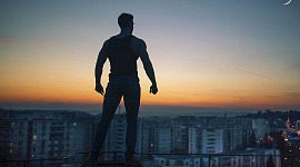 een silhouet van een man met gebalde vuisten die op een dak staat en uitkijkt over de stad