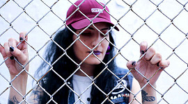 बेसबॉल टोपी पहने एक लड़की जंजीर की कड़ी बाड़ के पीछे खड़ी है