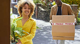 woman holding a potted plant, man holding a box that says Fragile, entering into a house
