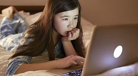una niña acostada en su cama usando una computadora portátil bajo el ojo de una cámara web