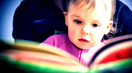 ایک بچہ اپنی ماں کی گود میں بیٹھا اور کتاب پڑھ رہا ہے۔