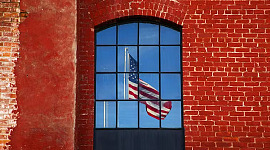 एक लाल ईंट की दीवार में एक खिड़की के माध्यम से देखा जाने वाला अमेरिकी झंडा