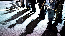 A line of police with riot shields on the street cast shadows onto the asphalt